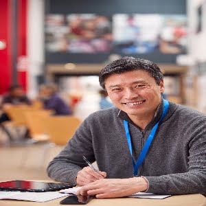 Portrait Of Mature Male Teacher Or Student With Digital Tablet Working At Table In College Hall